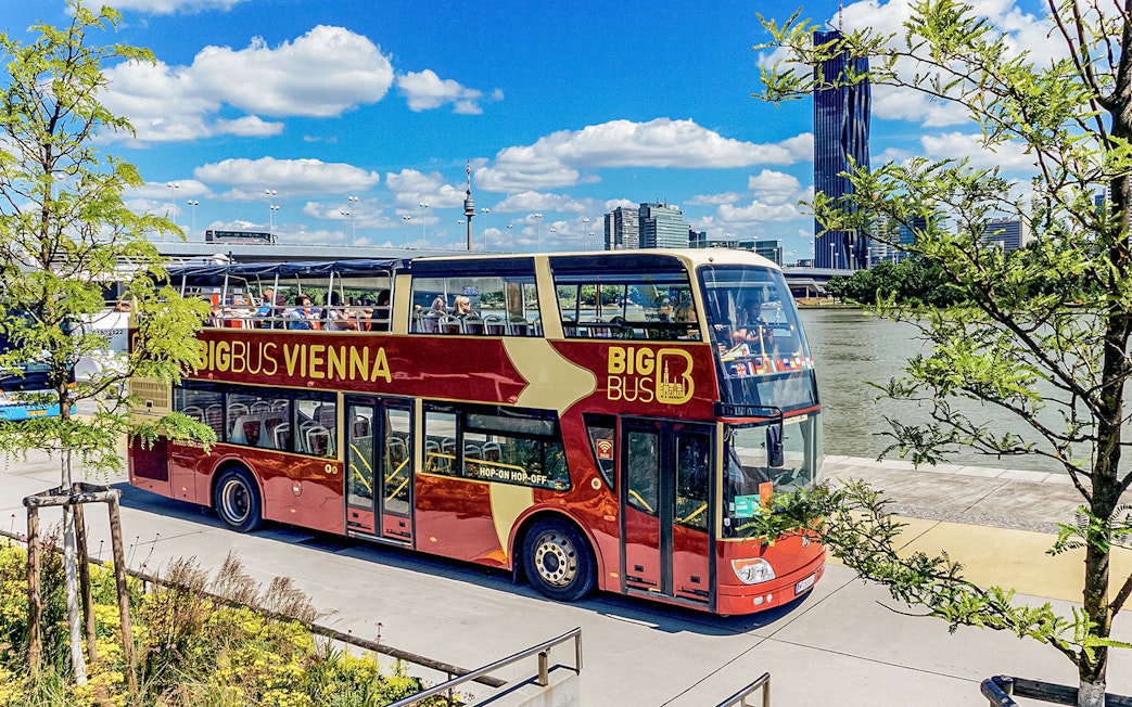 Tourists on Big Bus Vienna passing by the Danube River with city skyline in view.
