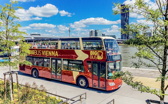 Tourists on Big Bus Vienna passing by the Danube River with city skyline in view.