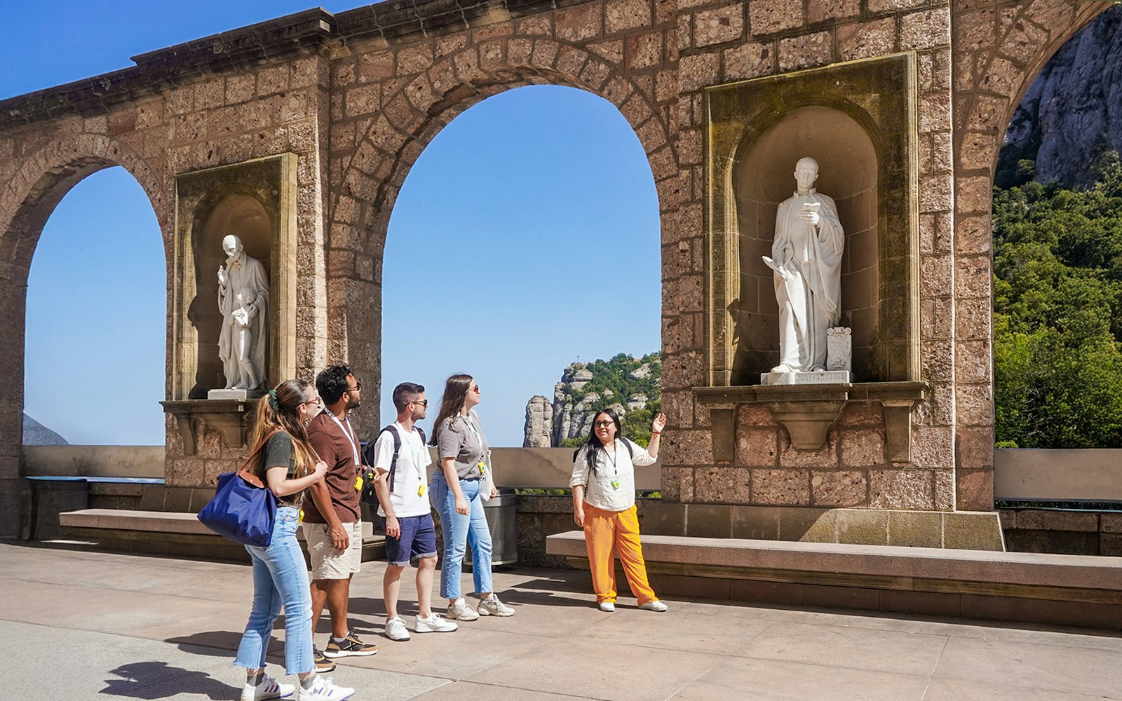 Tourists on a guided tour of Montserrat, viewing statues under stone arches.