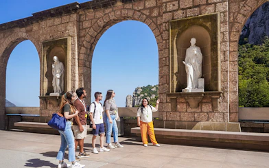 Tourists on a guided tour of Montserrat, viewing statues under stone arches.