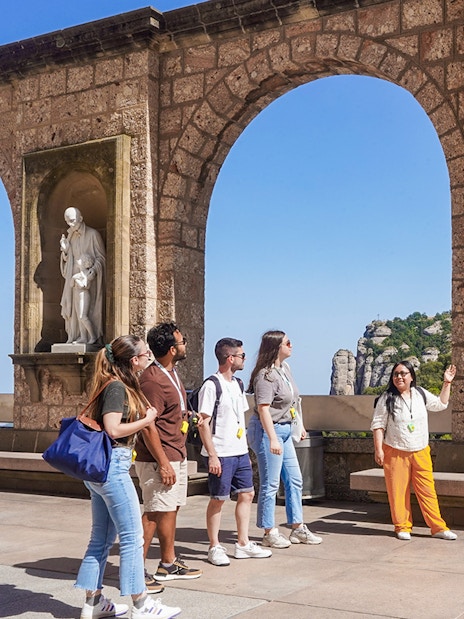 Tourists on a guided tour of Montserrat, viewing statues under stone arches.