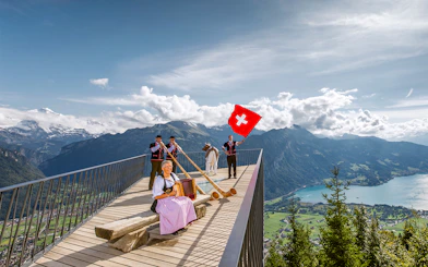 Swiss folk musicians performing at Herder Kulm with alphorns and Swiss flag, overlooking mountains.
