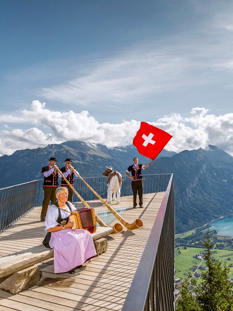 Swiss folk musicians performing at Herder Kulm with alphorns and Swiss flag, overlooking mountains.
