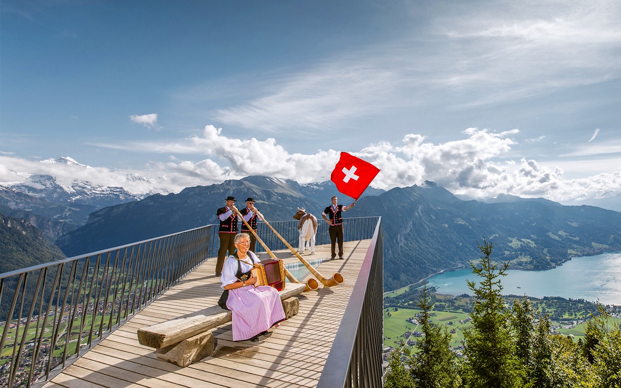 Swiss folk musicians performing at Herder Kulm with alphorns and Swiss flag, overlooking mountains.