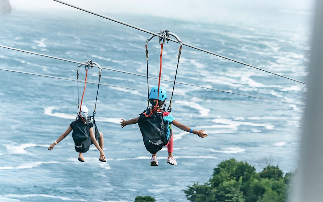 Two people ziplining over water towards a waterfall, wearing helmets and harnesses.