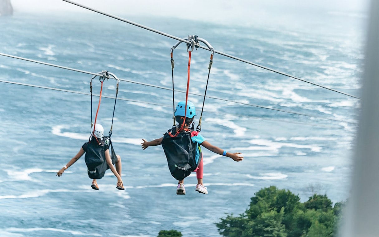 Two people ziplining over water towards a waterfall, wearing helmets and harnesses.