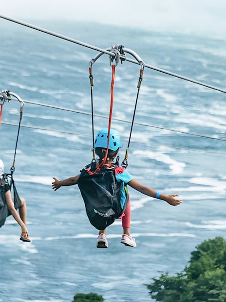 Two people ziplining over water towards a waterfall, wearing helmets and harnesses.