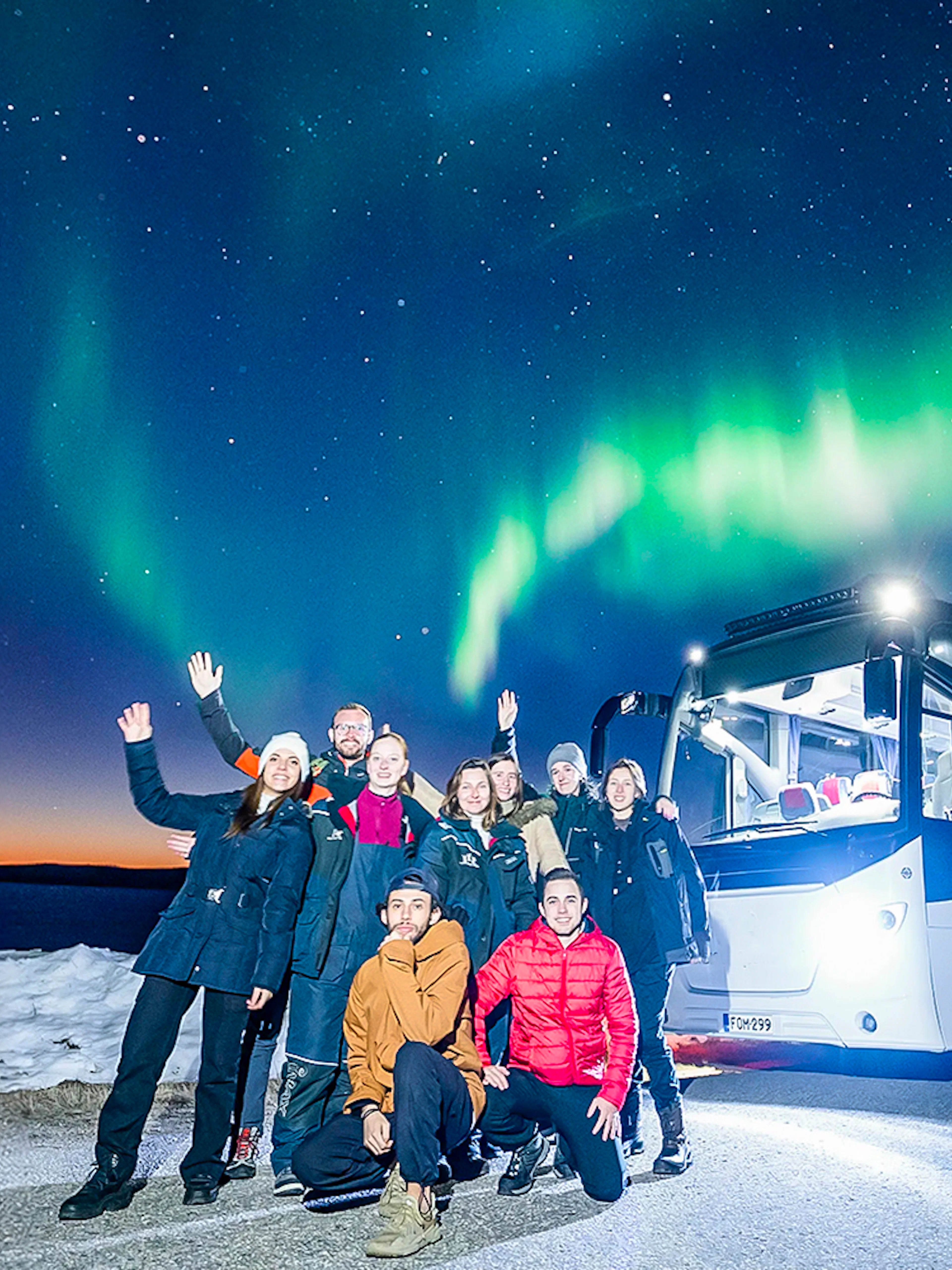 Tourists near a bus under Northern Lights in snowy landscape.