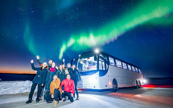 Tourists near a bus under Northern Lights in snowy landscape.