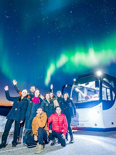 Tourists near a bus under Northern Lights in snowy landscape.