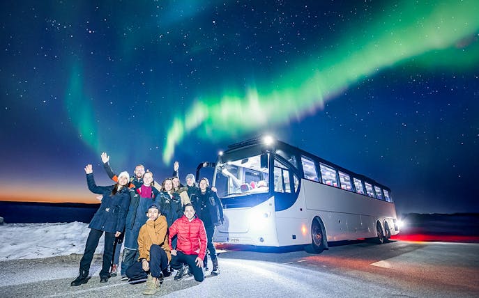 Tourists near a bus under Northern Lights in snowy landscape.