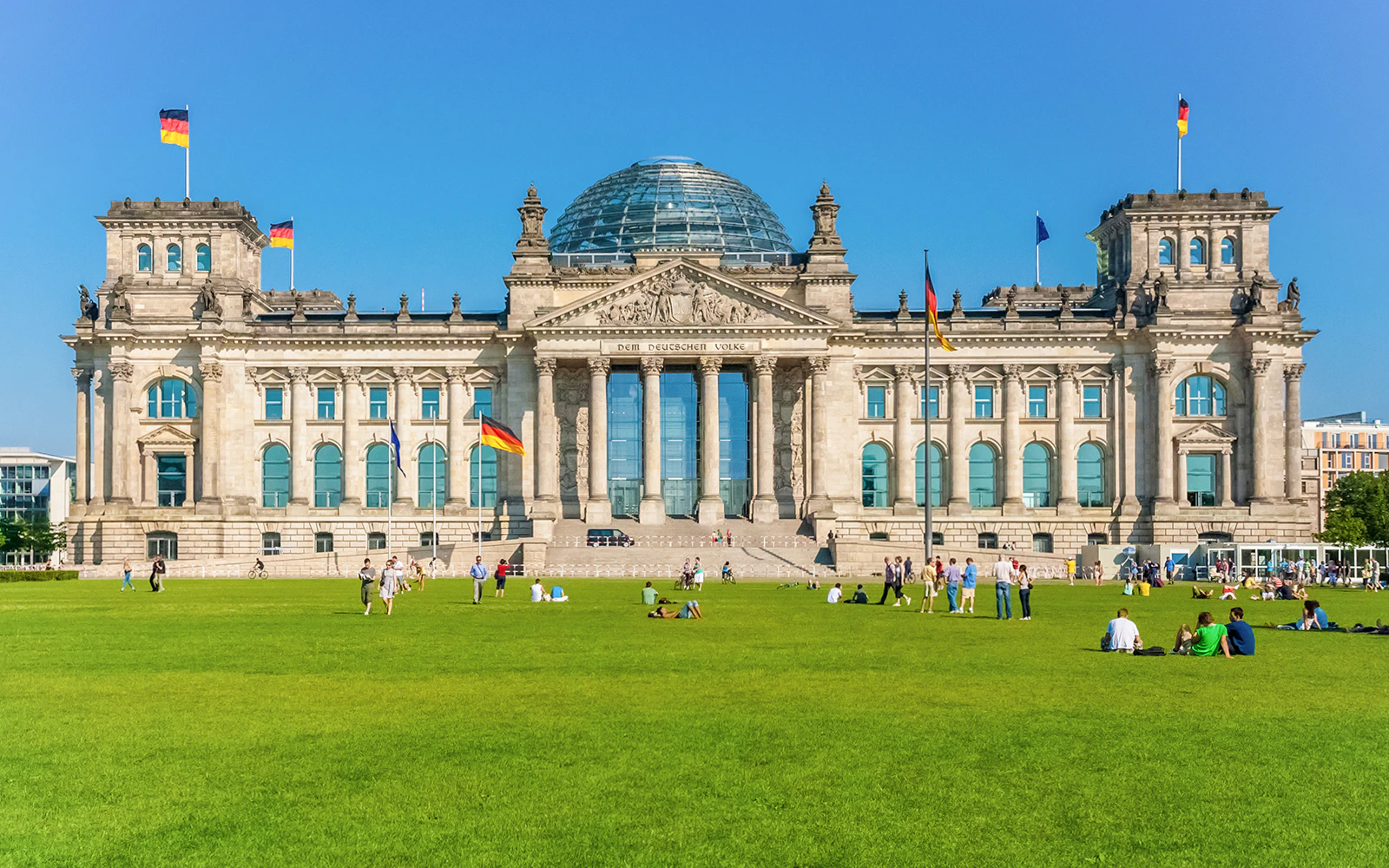 Reichstag Building in Berlin with people on the lawn, German flags flying.