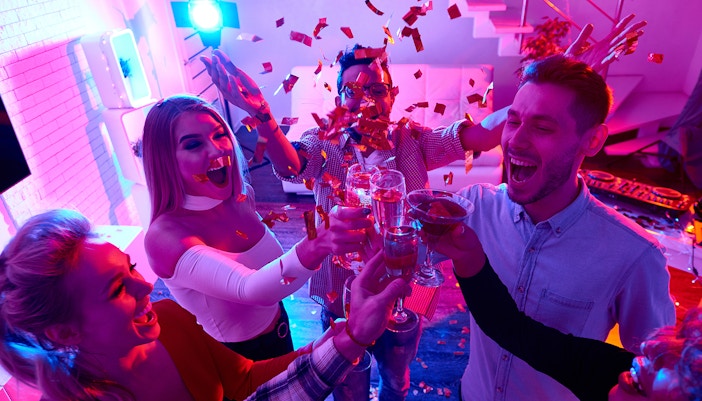 High angle view at group of happy friends enjoying holiday celebration at private house party raising glasses and toasting under bursts of confetti