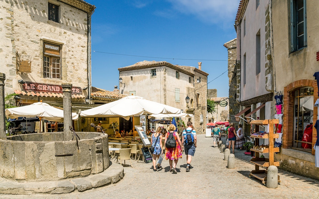 People walking through a historic street in Carcassonne, France, near a stone well and outdoor café.