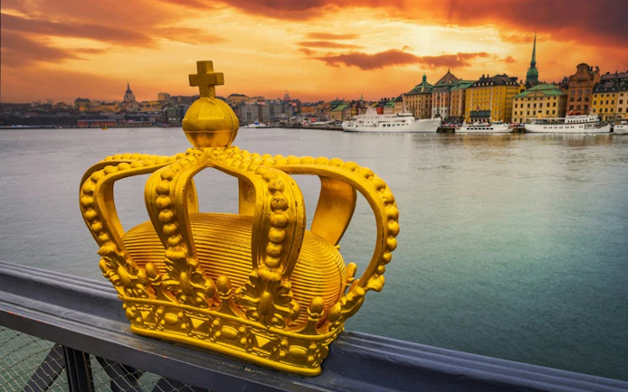 Golden crown on Stockholm bridge with cityscape and sunset in background.