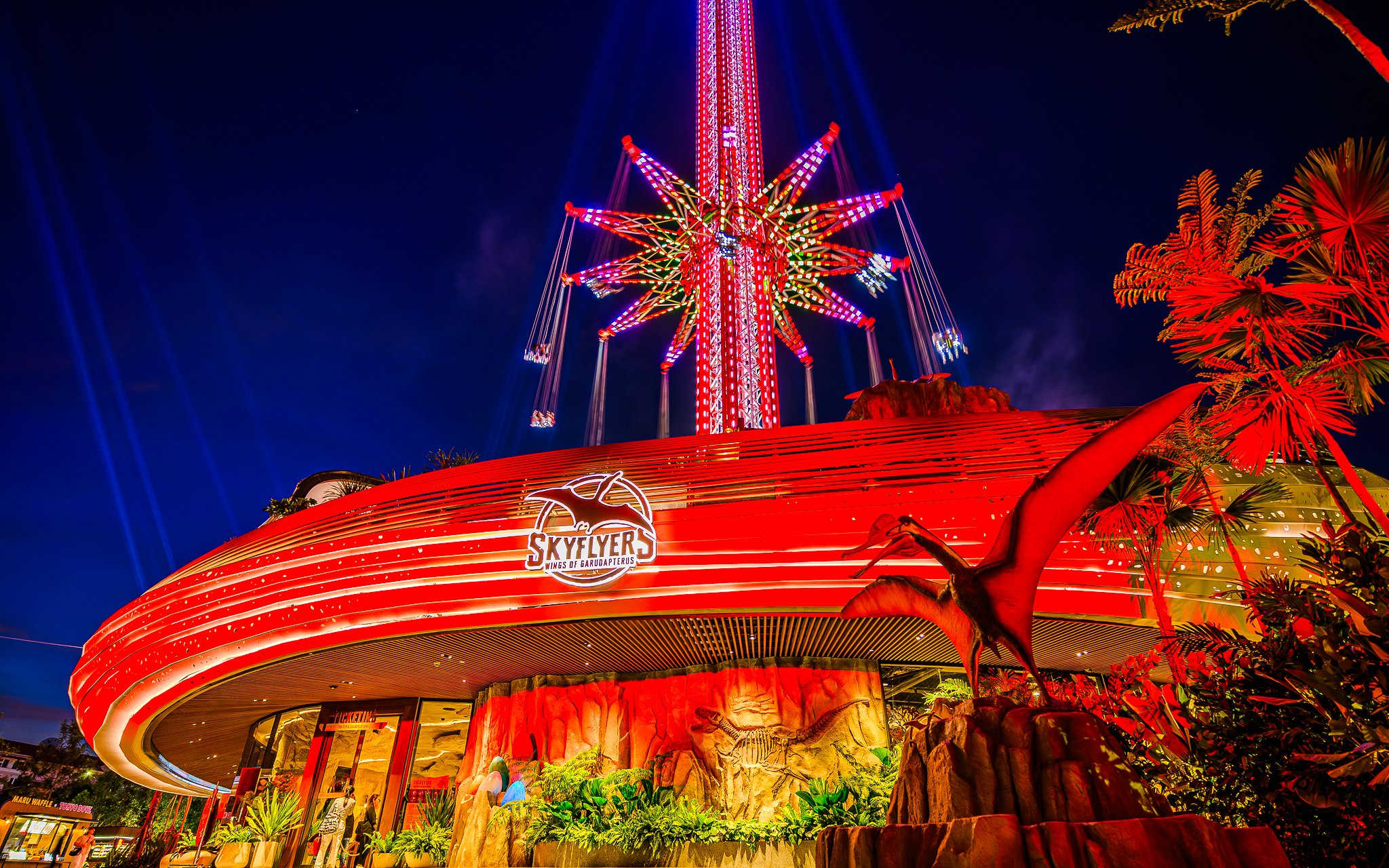 SkyFlyers ride illuminated at night in Asiatique Bangkok, featuring vibrant lights and dinosaur decor.