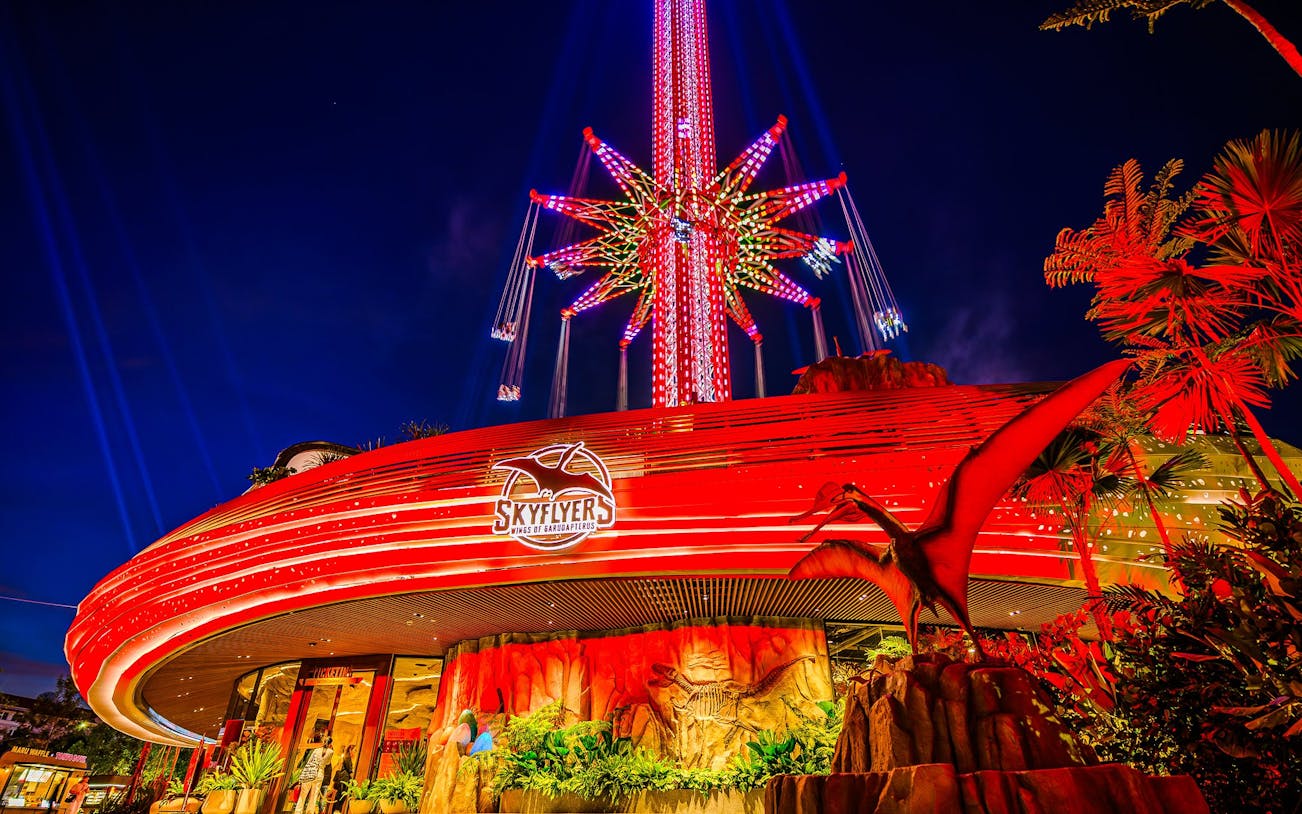 SkyFlyers ride illuminated at night in Asiatique Bangkok, featuring vibrant lights and dinosaur decor.