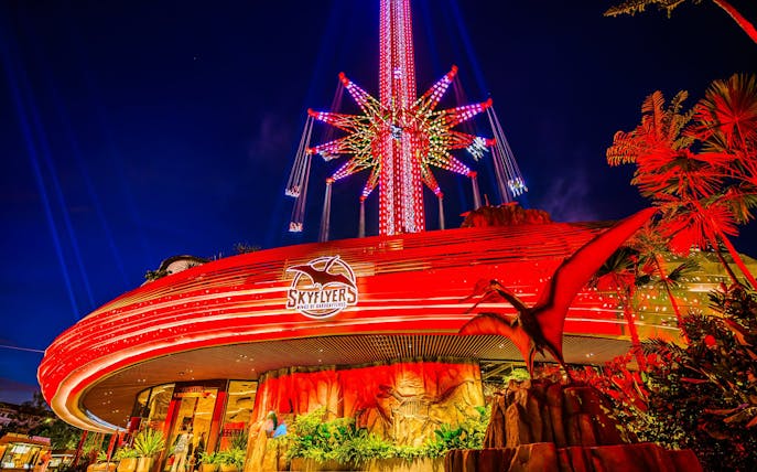 SkyFlyers ride illuminated at night in Asiatique Bangkok, featuring vibrant lights and dinosaur decor.