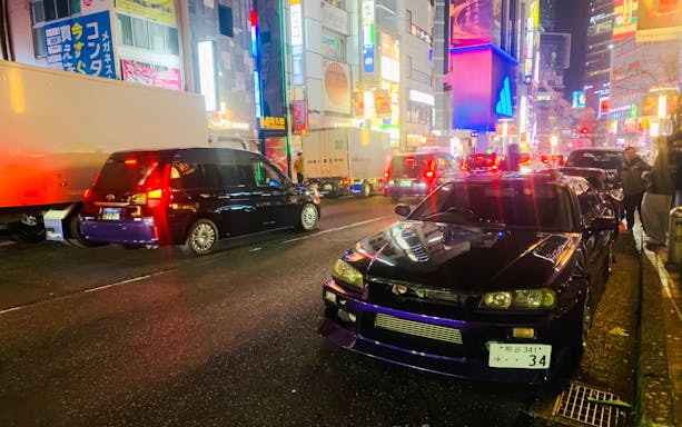 Supercar parked on a busy street in Tokyo at night, surrounded by vibrant city lights.