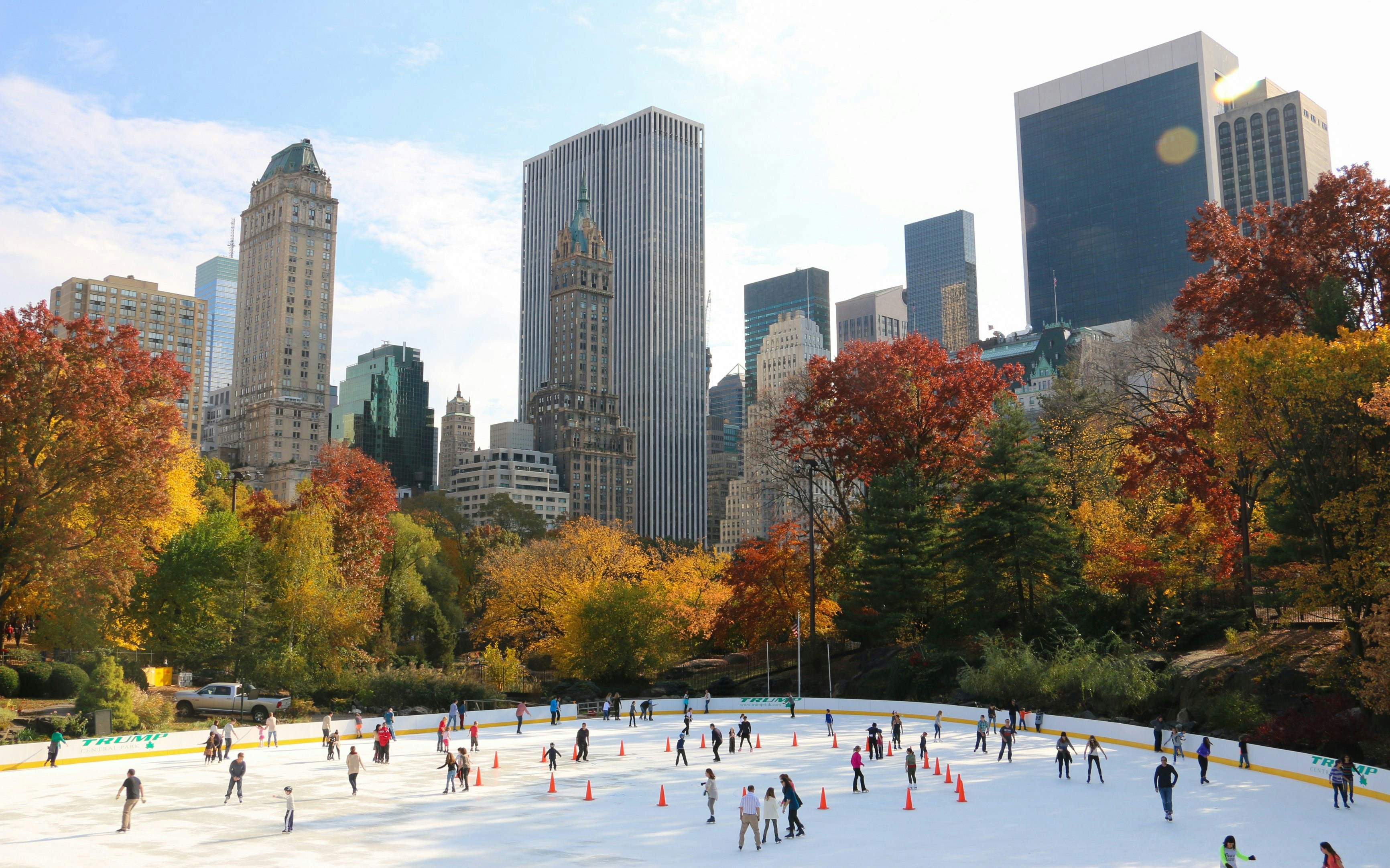 Ice skaters at Wollman Rink in Central Park, New York, with autumn trees and city skyline.