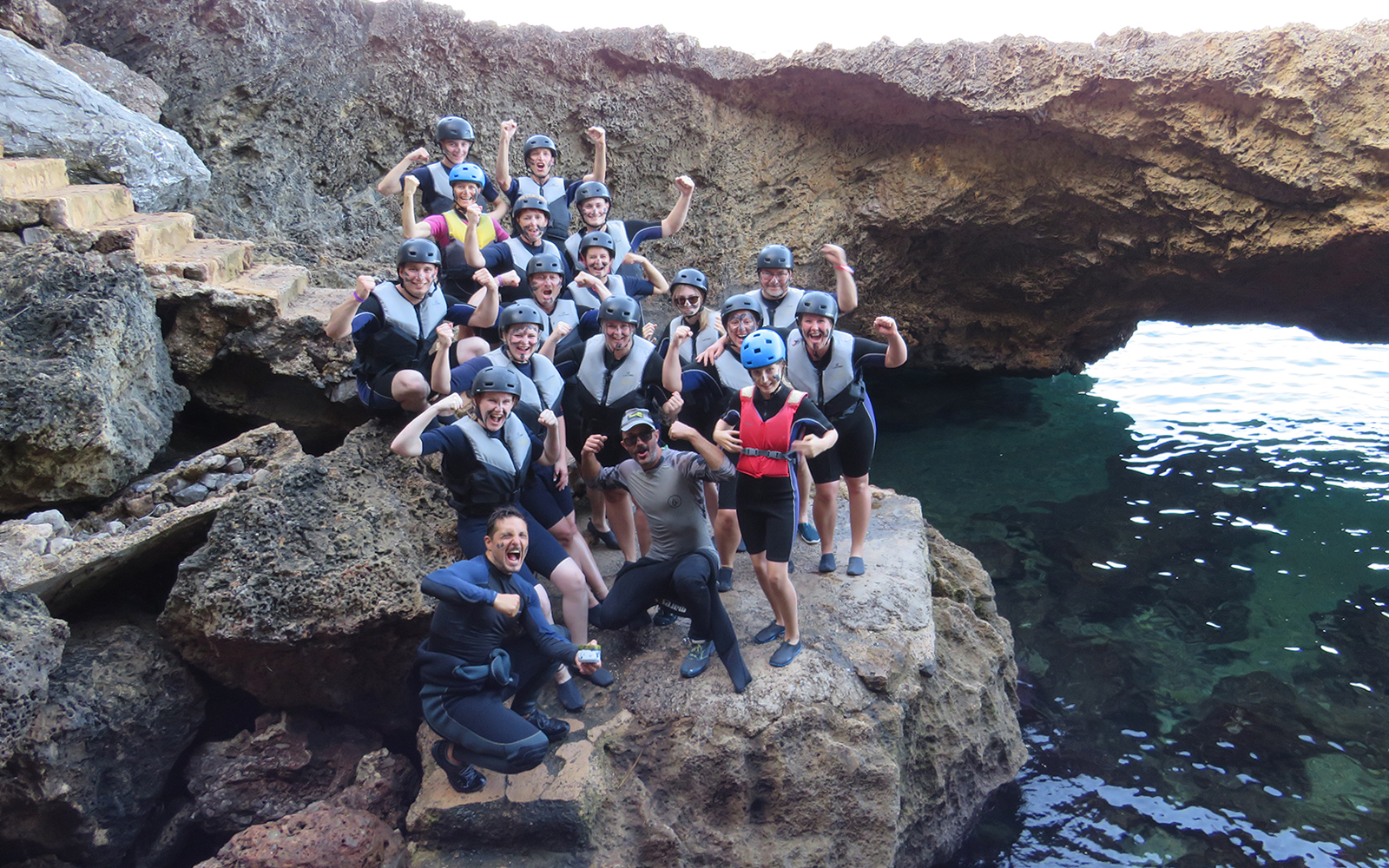 Group of people in helmets and life vests posing on rocky shore near sea cave in Mallorca.