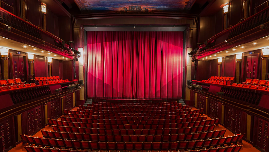 Interior of a London theatre with red seats and closed stage curtain.