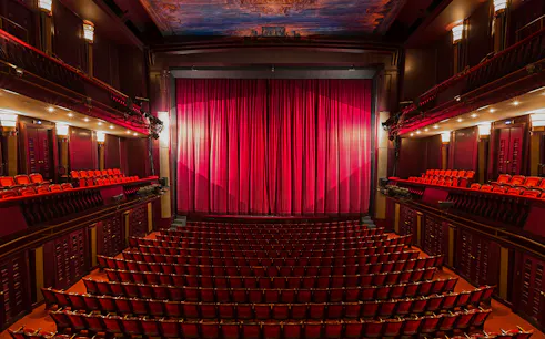 Interior of a London theatre with red seats and closed stage curtain.