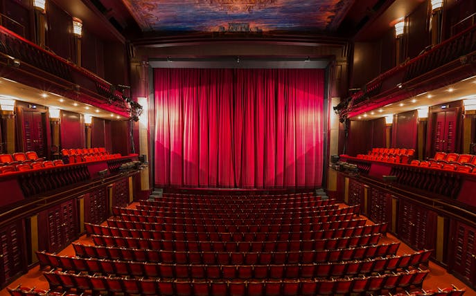 Interior of a London theatre with red seats and closed stage curtain.