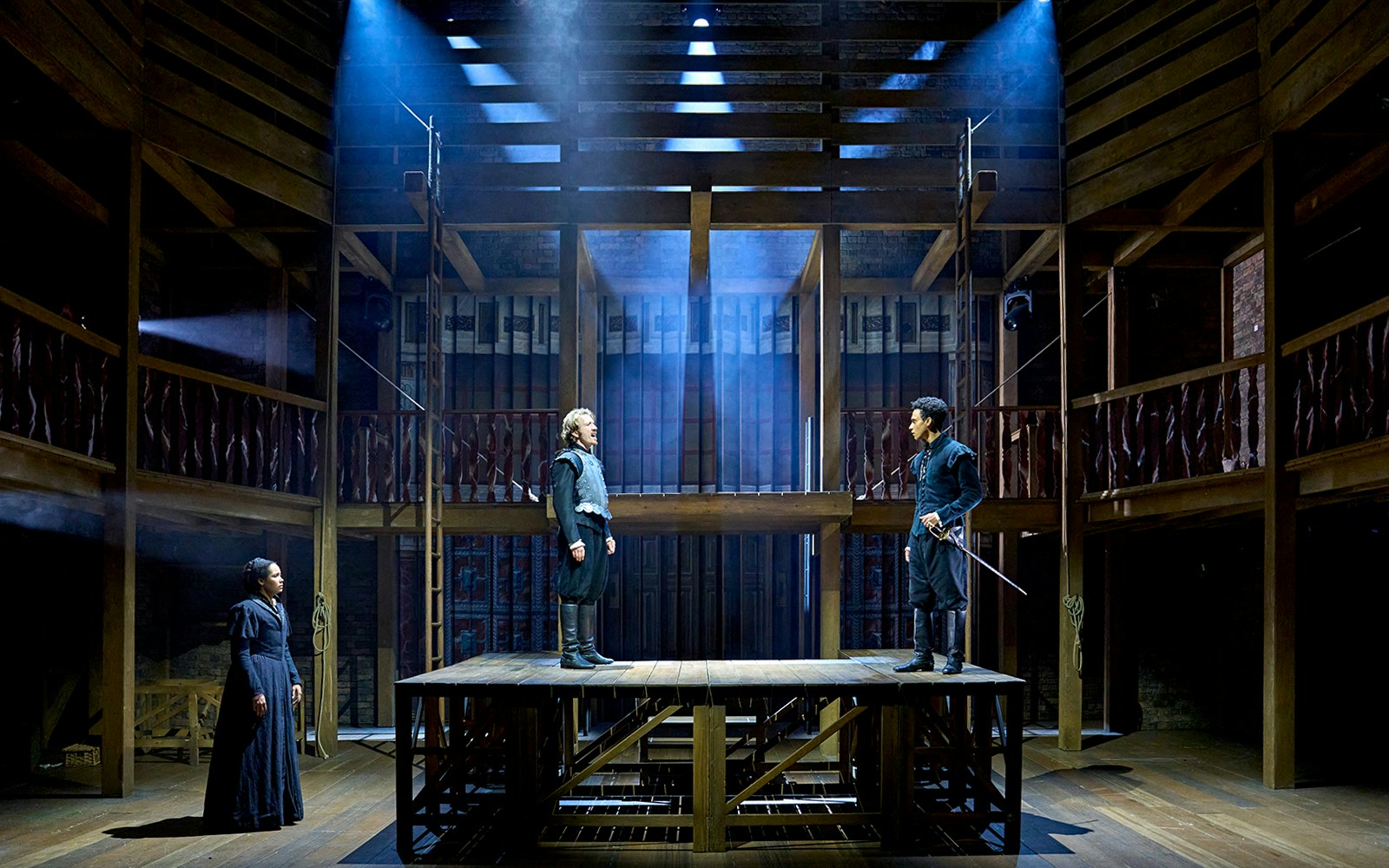 Actors performing a scene from "Hamnet" on a wooden stage with dramatic lighting.