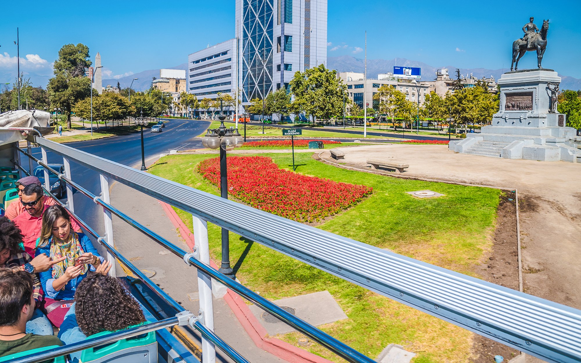 Santiago hop-on hop-off bus passing Plaza Baquedano with cityscape and statue.