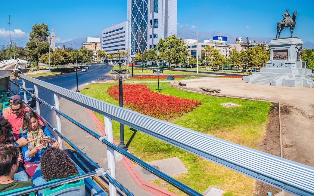 Santiago hop-on hop-off bus passing Plaza Baquedano with cityscape and statue.