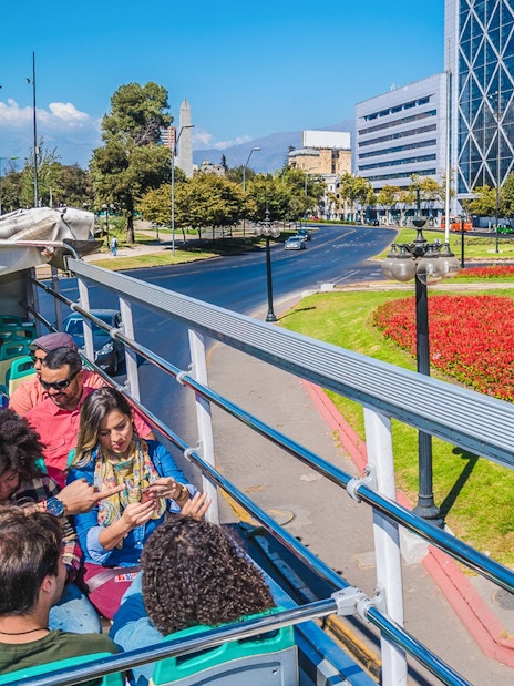 Santiago hop-on hop-off bus passing Plaza Baquedano with cityscape and statue.