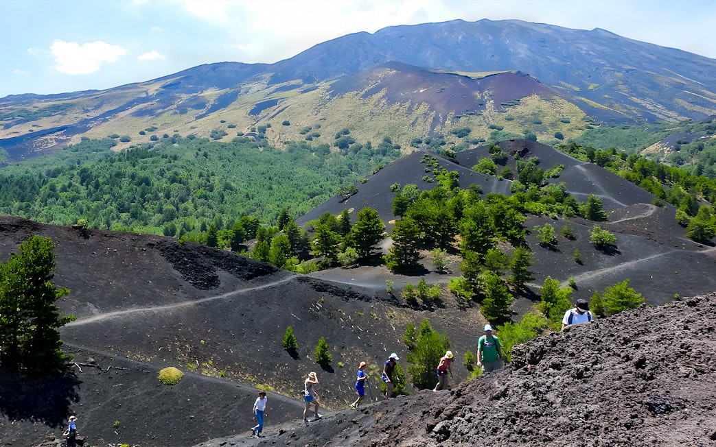 Hikers exploring volcanic terrain on Mount Etna with lush greenery in the background.