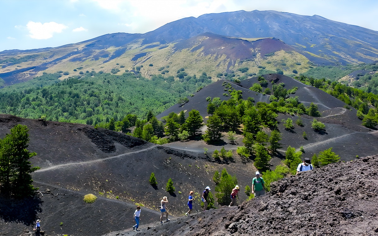 Hikers exploring volcanic terrain on Mount Etna with lush greenery in the background.