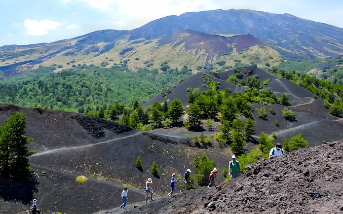 Hikers exploring volcanic terrain on Mount Etna with lush greenery in the background.