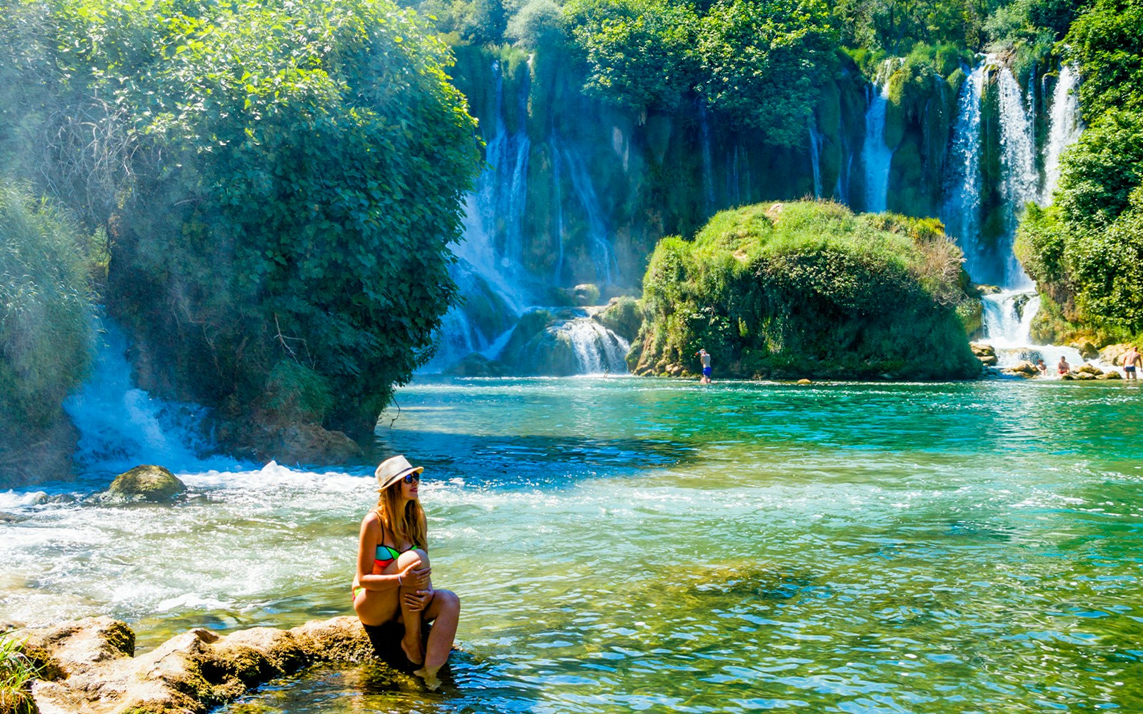 Tourist sitting by Kravica Waterfalls, Bosnia and Herzegovina, with lush greenery.