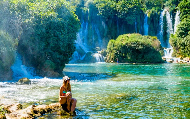 Tourist sitting by Kravica Waterfalls, Bosnia and Herzegovina, with lush greenery.