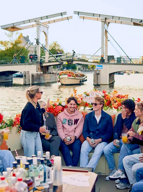 People socializing on a flower-adorned boat in Amsterdam canal with bridge in background.