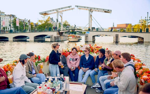 People socializing on a flower-adorned boat in Amsterdam canal with bridge in background.