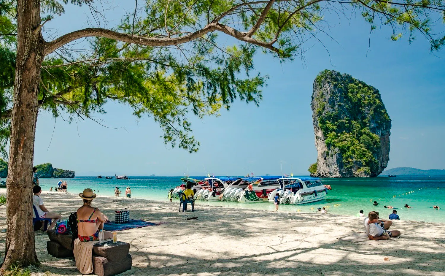 Longtail boats docked on a sandy beach with tourists relaxing and snorkeling near a limestone island in Krabi.