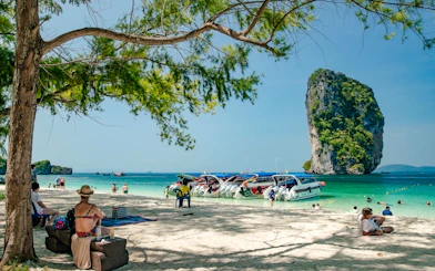 Longtail boats docked on a sandy beach with tourists relaxing and snorkeling near a limestone island in Krabi.