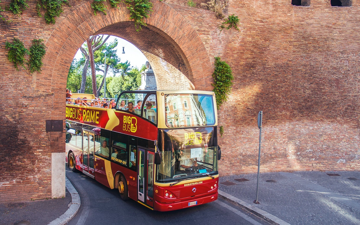 Hop-on hop-off bus with tourists passing through ancient Roman arch.