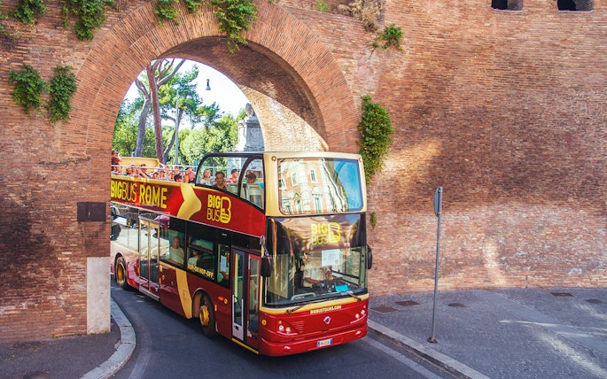 Hop-on hop-off bus with tourists passing through ancient Roman arch.