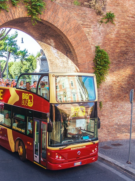 Hop-on hop-off bus with tourists passing through ancient Roman arch.