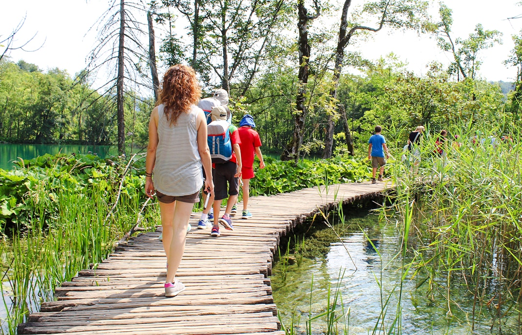 Tourists walking on a wooden bridge at Plitvice Lakes, Croatia surrounded by lush greenery.