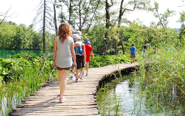 Tourists walking on a wooden bridge at Plitvice Lakes, Croatia surrounded by lush greenery.