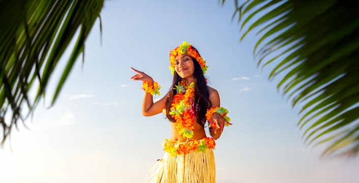 Hula dancer performing by the ocean with palm leaves in the foreground.
