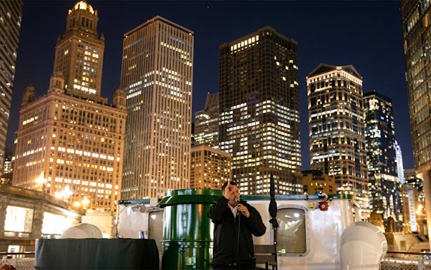 Tour guide narrating on Chicago Haunted River "Creepy Cruise" with city skyline at night.