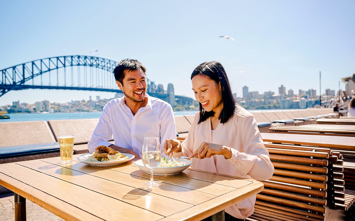 Couple dining outdoors with Sydney Opera House and Harbour Bridge in the background.