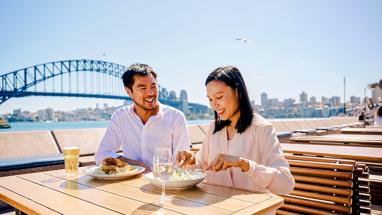 Couple dining outdoors with Sydney Opera House and Harbour Bridge in the background.